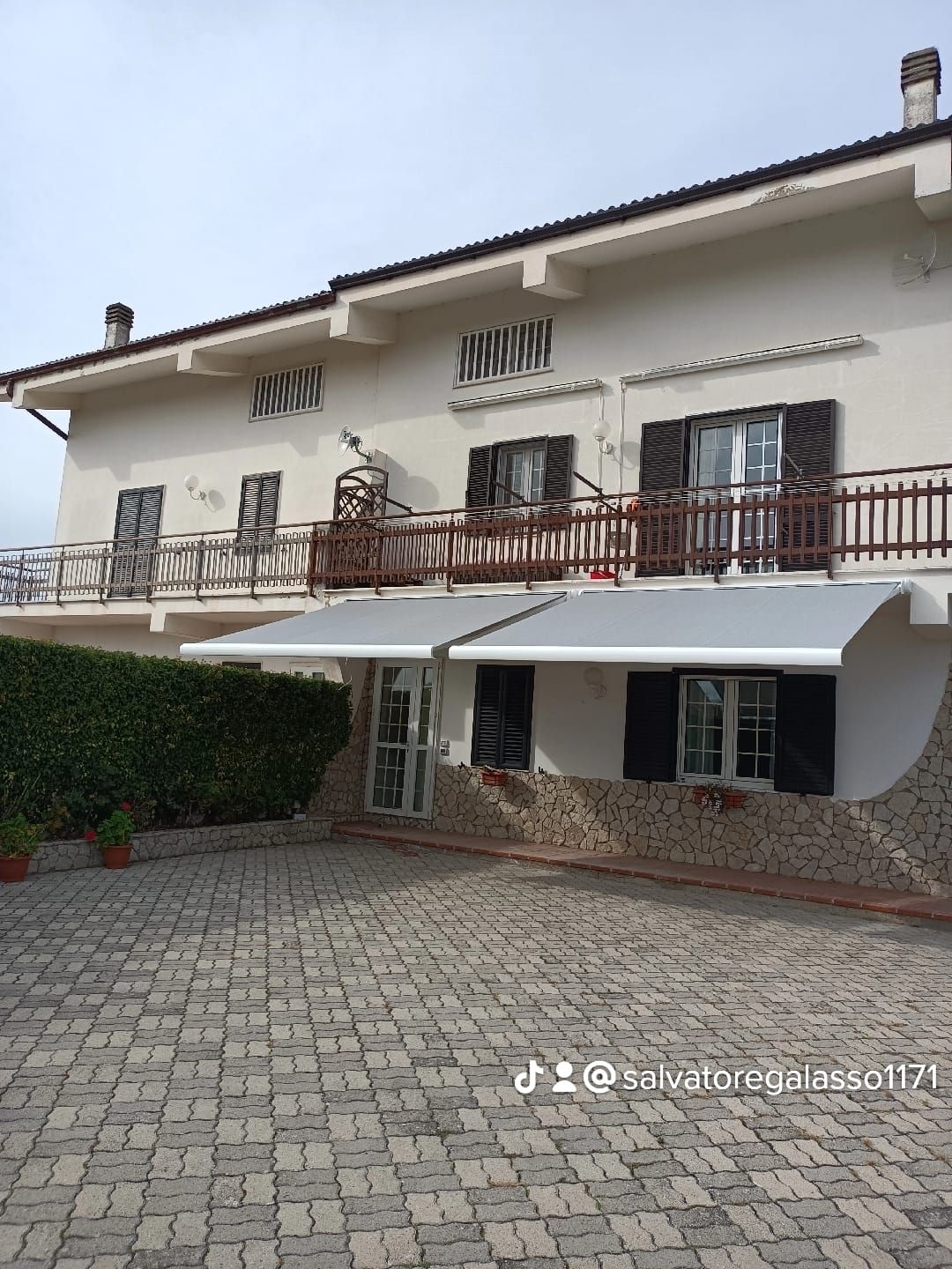 Casa a due piani con facciata bianca, balcone, tende da sole e vialetto d'accesso pavimentato nel cortile.