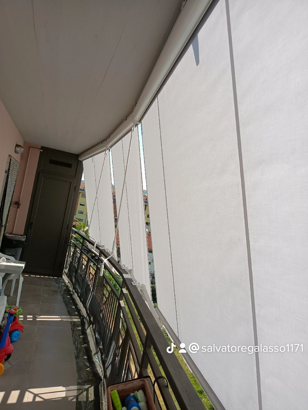 Balcone coperto con ringhiera bianca, piante in vaso e uno stretto passaggio che si affaccia su un'area esterna.