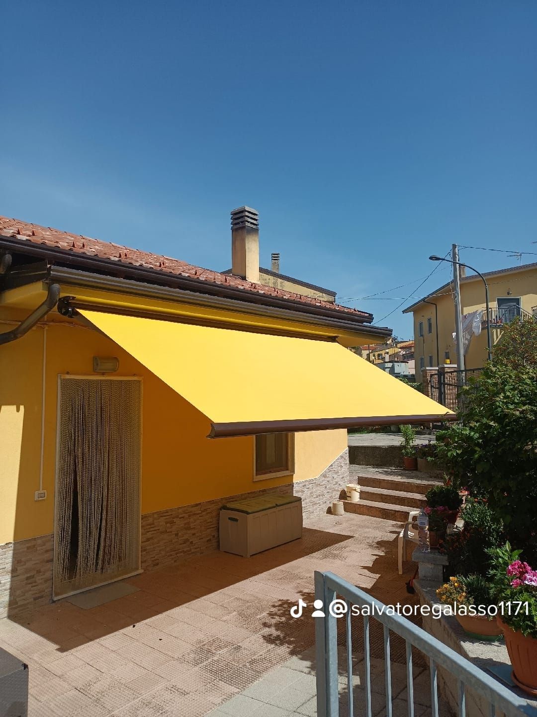 Casa gialla con un'ampia tenda da sole, scalini in pietra e un cielo azzurro limpido in un cortile soleggiato