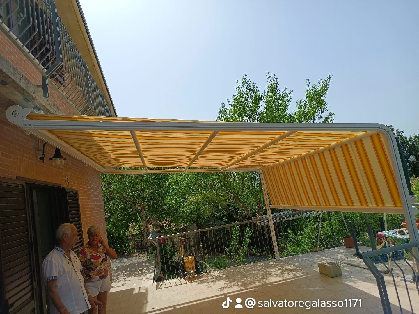 Tenda a strisce gialle attaccata a una casa in mattoni, che ripara due persone su un patio con alberi sullo sfondo.