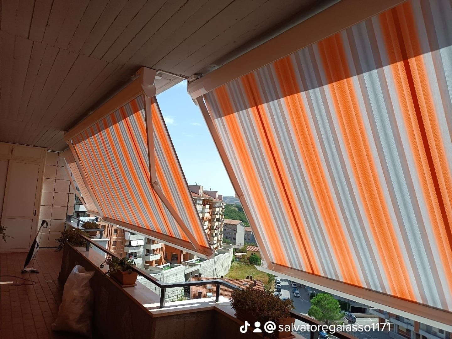 Balcone con tende a righe arancioni e bianche che si affaccia su edifici e cielo azzurro.