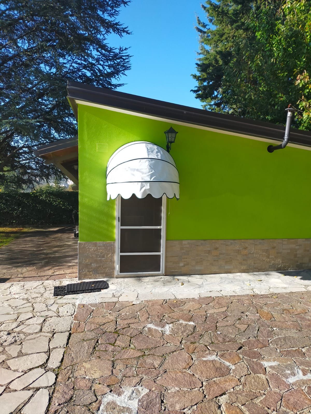 Una parete di un edificio di un verde brillante con una piccola tenda da sole bianca sopra una porta, accanto a un patio in pietra e ad alcuni alberi.