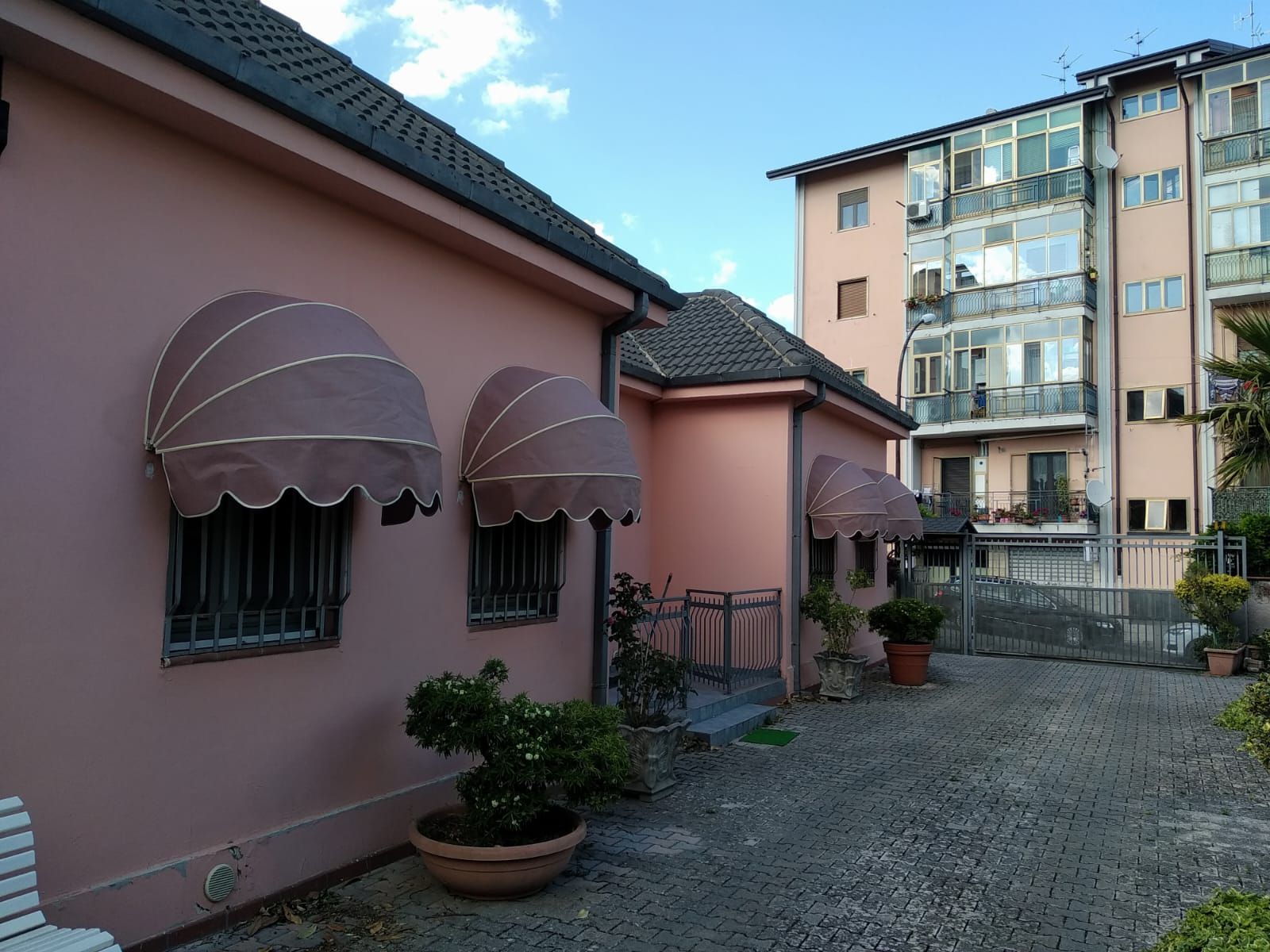 Esterno di una casa rosa con tende da sole, piante in vaso e un cortile adiacente a un condominio.