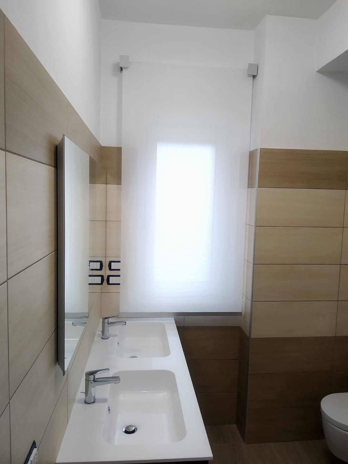 Bagno moderno con doppio lavabo, ampio specchio, pareti rivestite con piastrelle beige e finestra satinata.