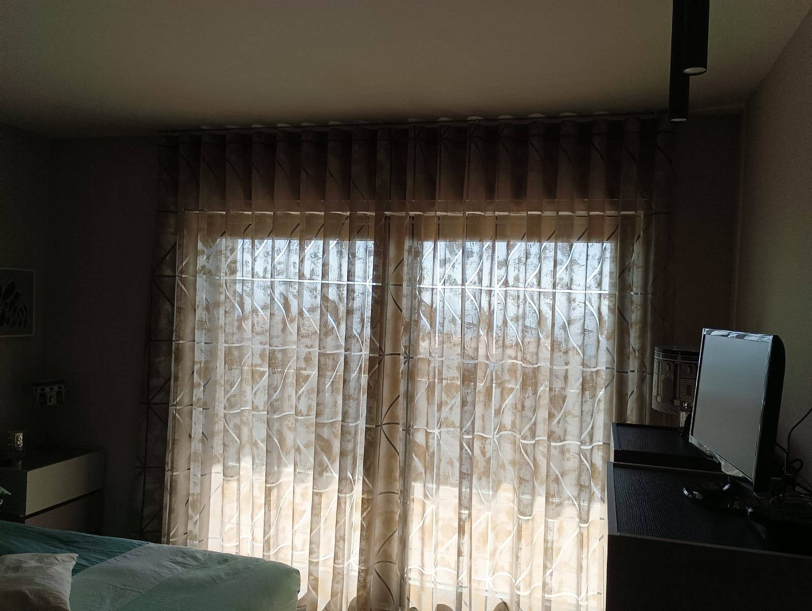 Camera da letto in penombra con tende trasparenti che brillano davanti a una finestra e un televisore su una cassettiera.
