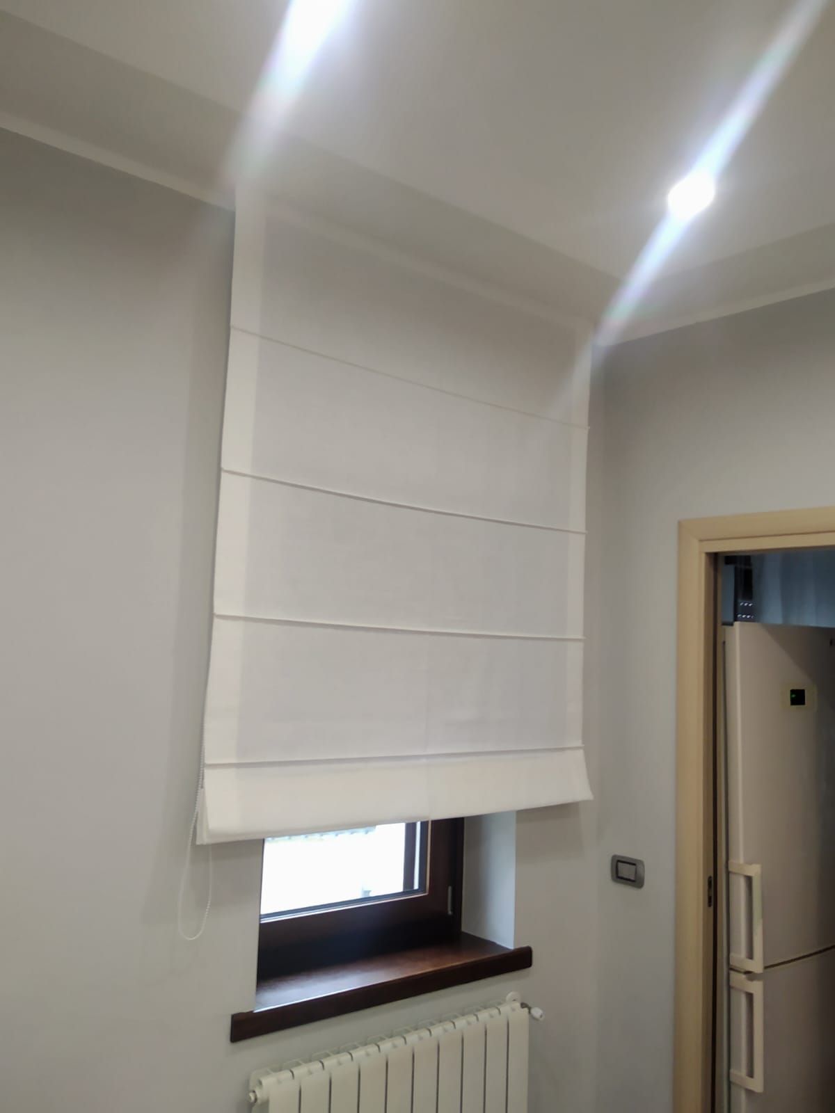 Tenda a pacchetto bianca che copre una piccola finestra in una stanza grigio chiaro accanto a una porta.