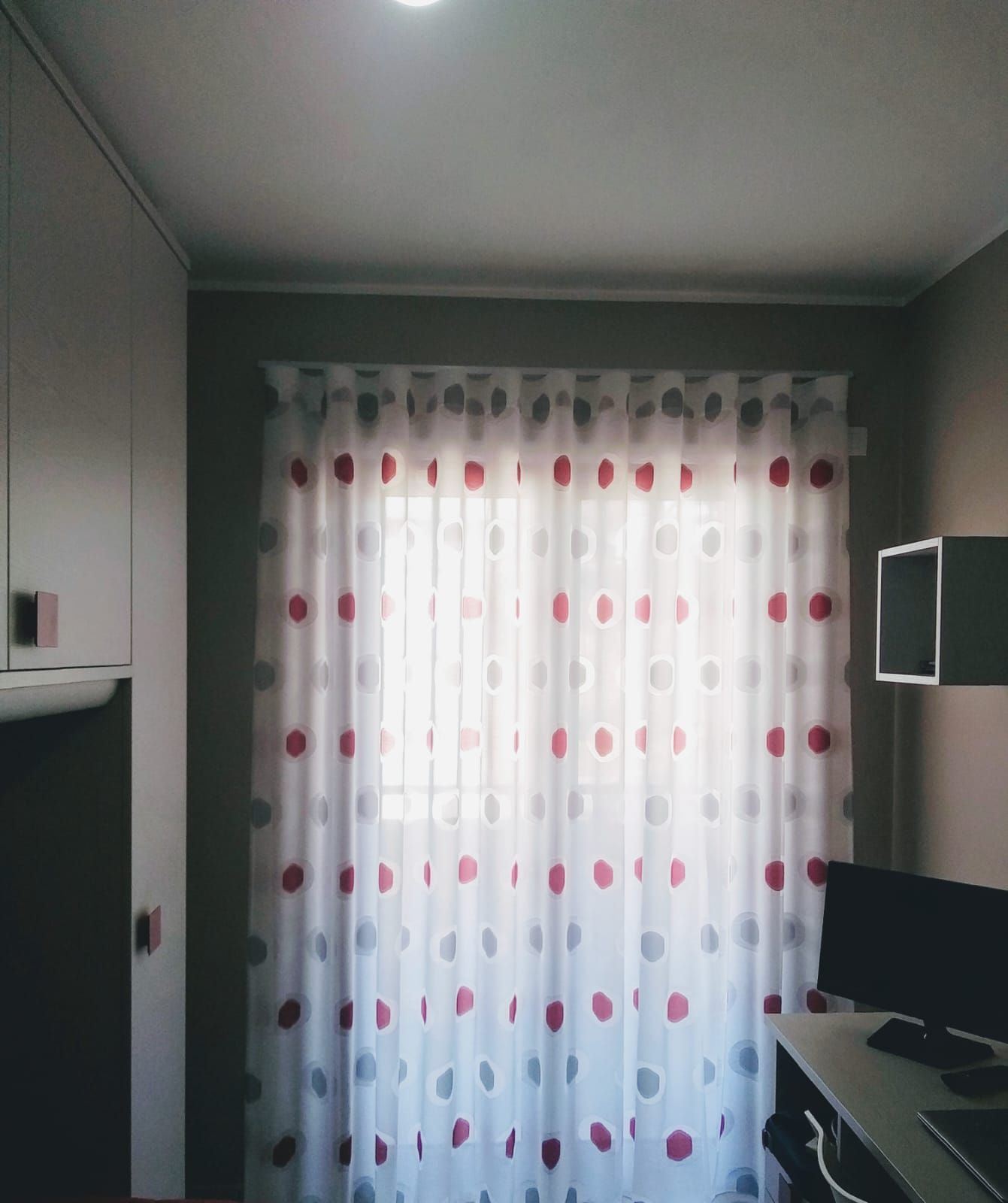 Piccola stanza con tende bianche, scrivania e monitor, e armadi a muro sulla sinistra.