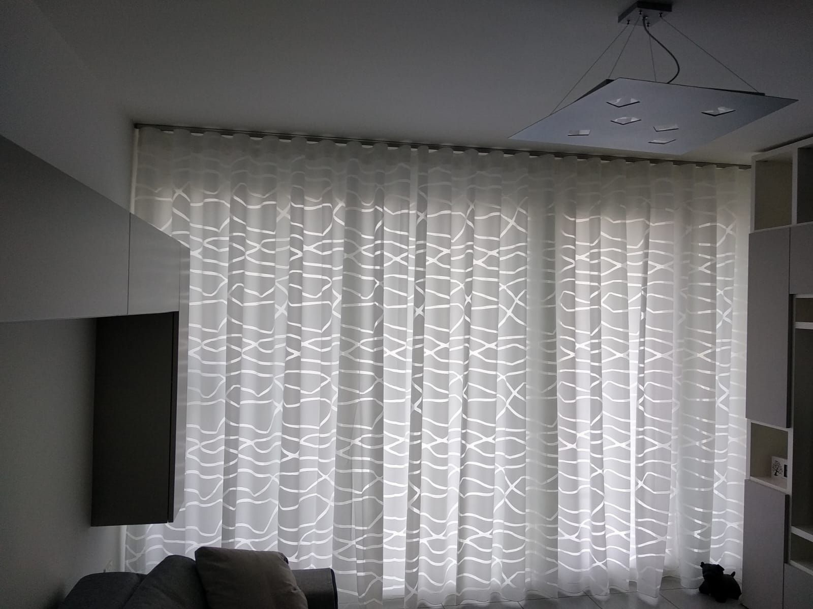 Luminoso soggiorno con tende bianche che coprono un'ampia finestra e un televisore a parete sulla sinistra.