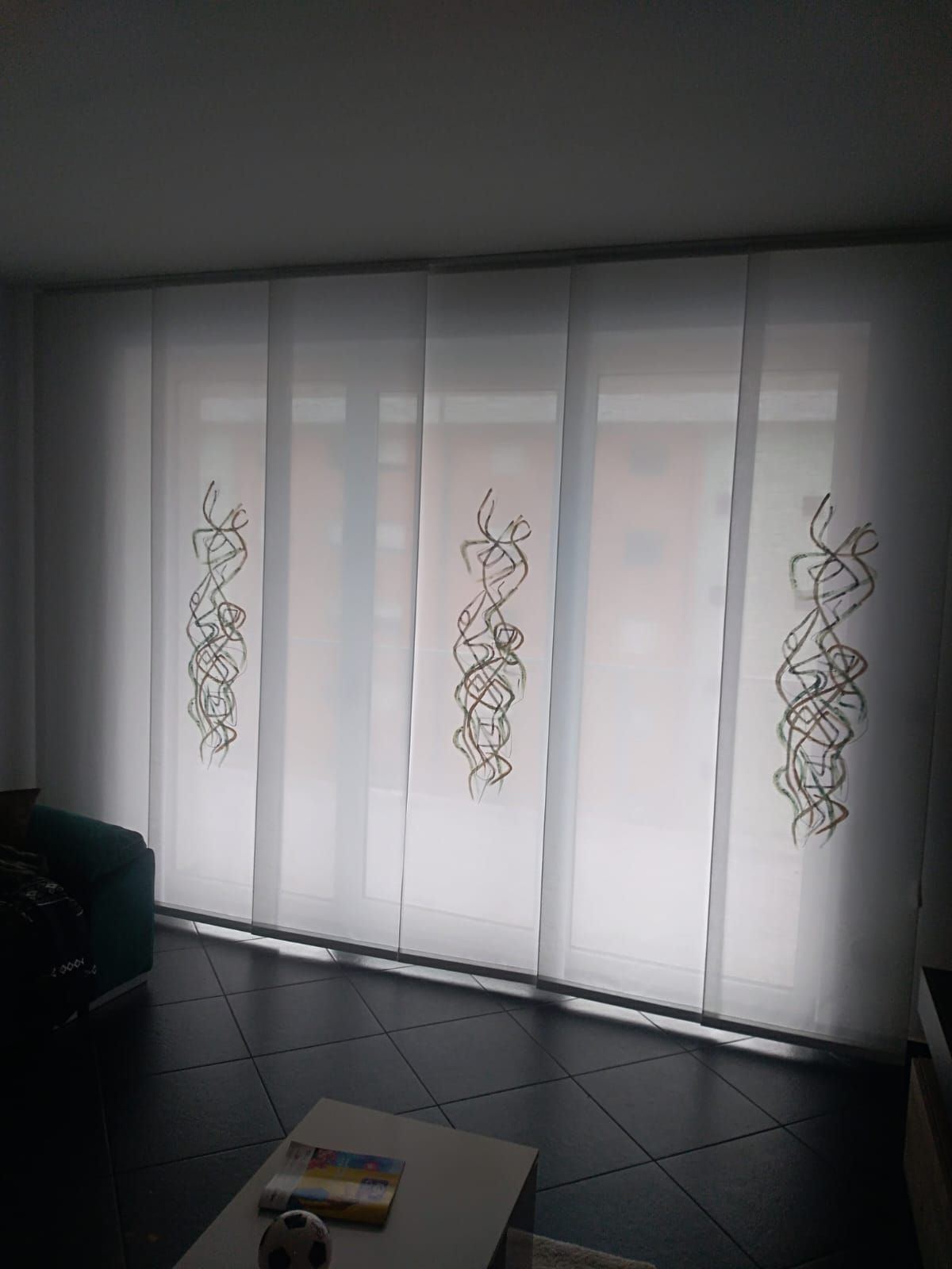 Tende bianche trasparenti con ricami floreali in una stanza poco illuminata, con un tavolino in primo piano.