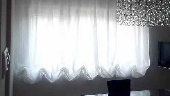 Tende bianche trasparenti che brillano alla luce del giorno, con un piccolo televisore scuro sulla destra.