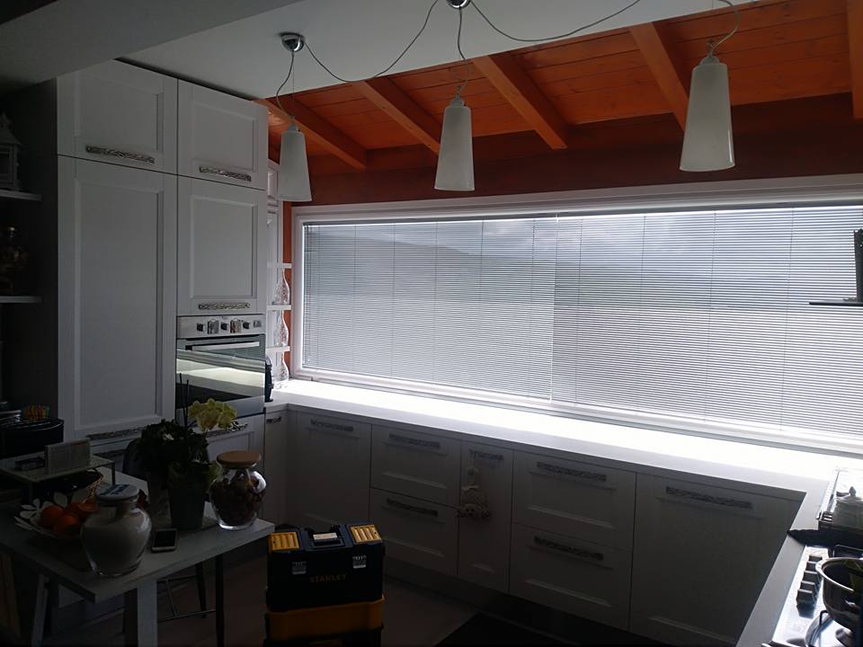 Cucina luminosa con mobili bianchi, grandi tende alle finestre, lampade a sospensione e travi in legno a vista sul soffitto.