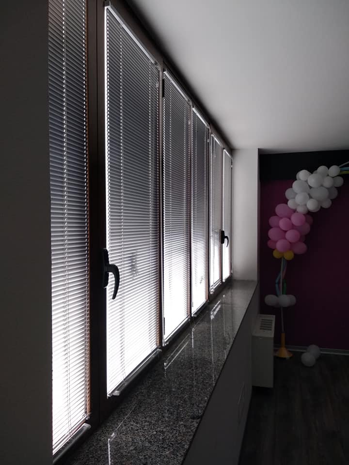 Corridoio d'ufficio poco illuminato con alte finestre e persiane; decorazione di palloncini rosa e bianchi in fondo.