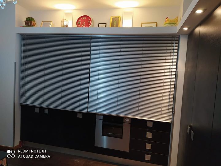 Parete cucina moderna con persiane argentate chiuse, mobili neri e piano di lavoro bianco.