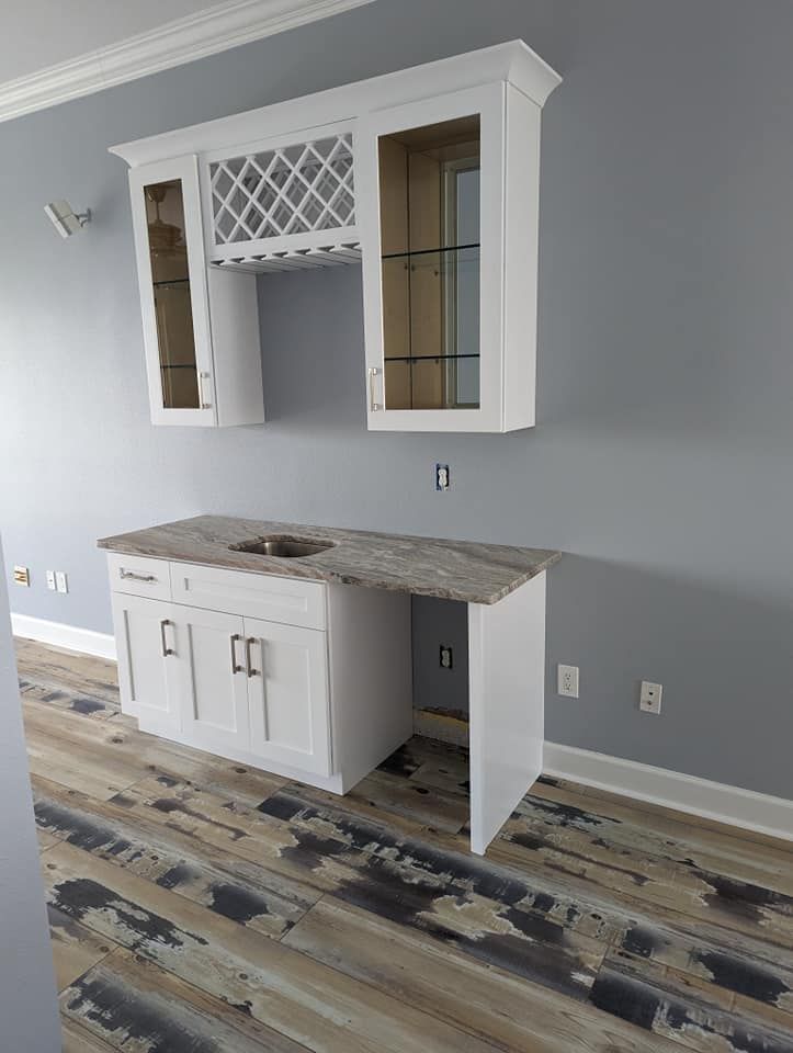 White bar cabinet with granite countertop and overhead shelving on gray wall, over wood floor.