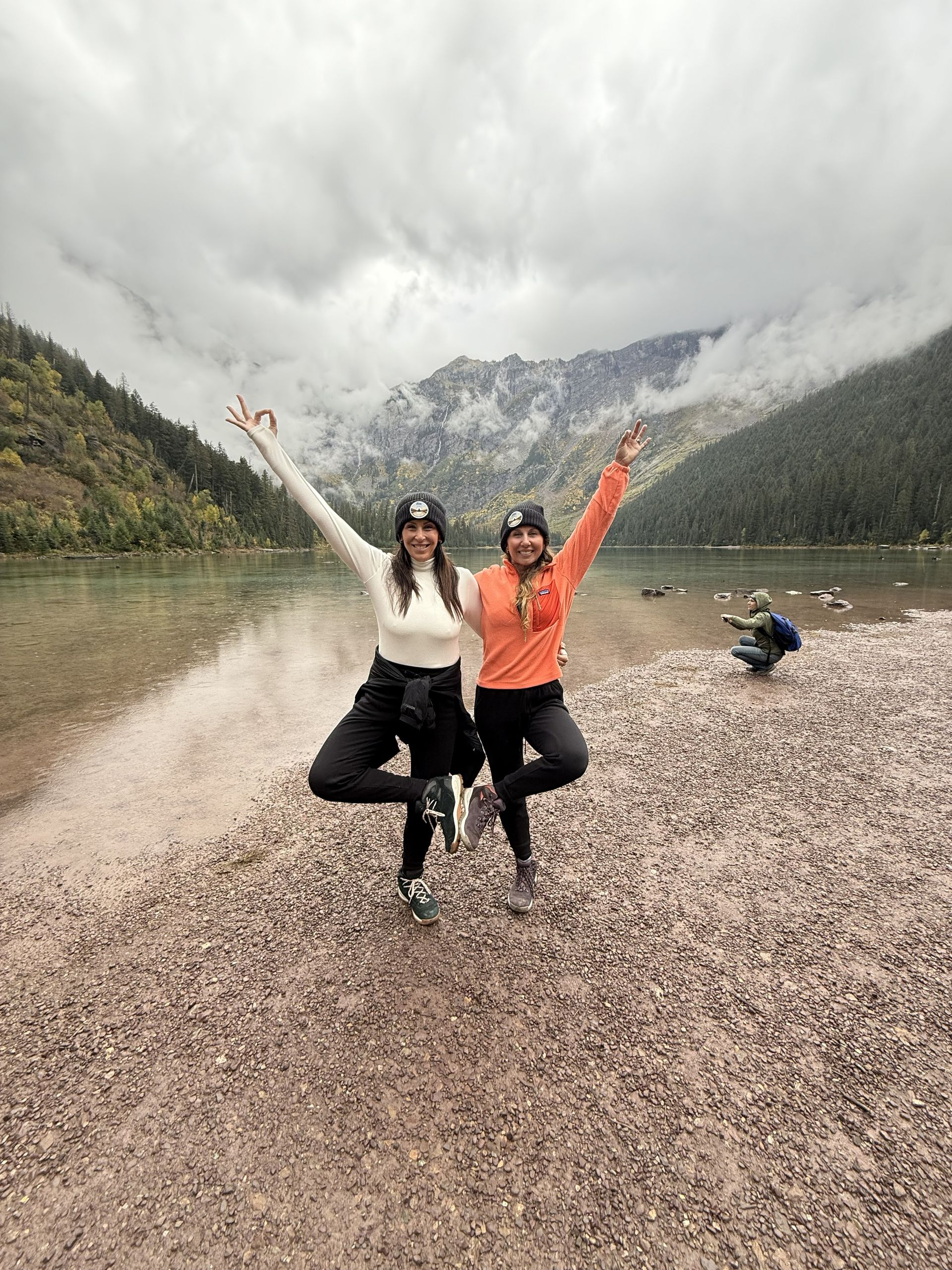 Two people posing with raised arms by a lake and mountains under cloudy skies, with a dog in the background.