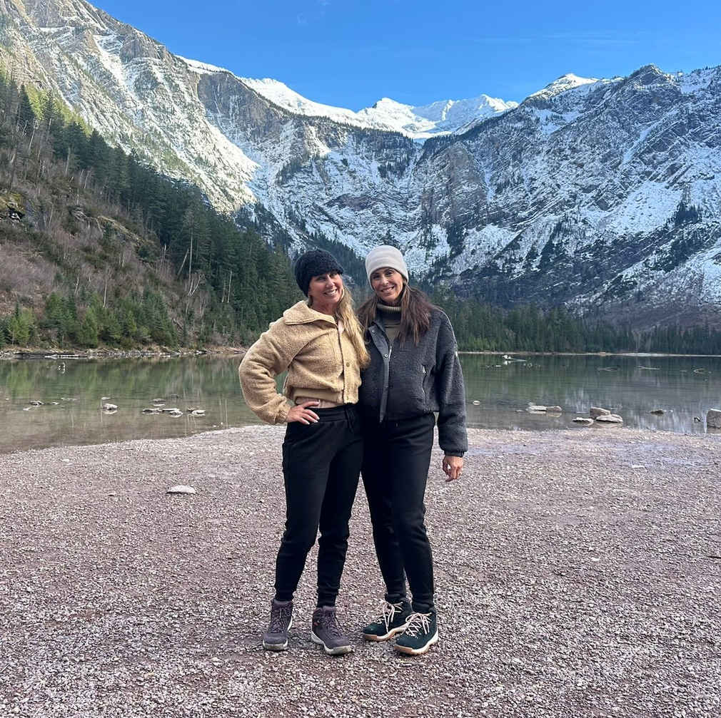 Two people standing on a rocky lakeshore with snowy mountains in the background