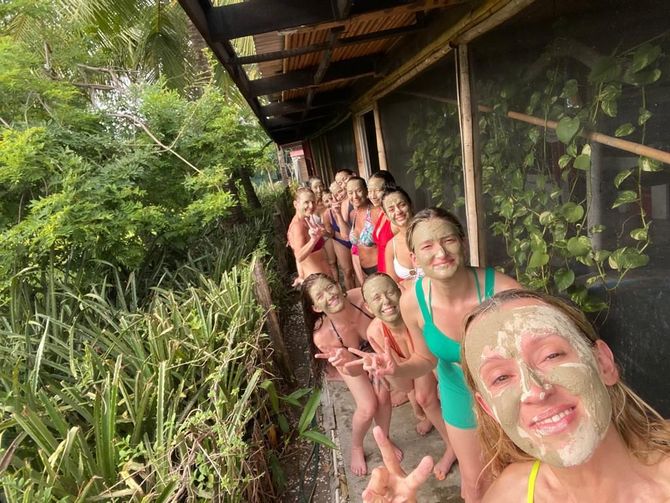 People in towels pose along a wooden walkway beside lush greenery, smiling with mud masks on their faces.