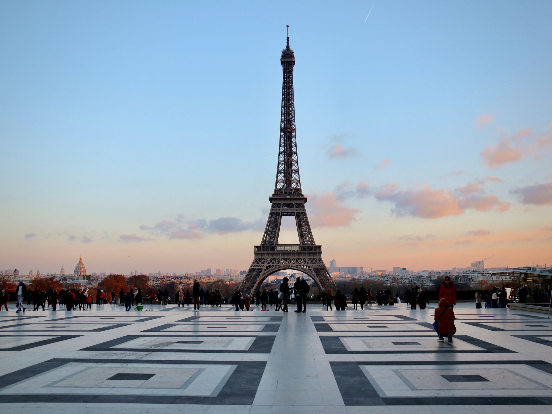 Eiffel Tower at sunset with people gathered on a patterned plaza under a pink-blue sky