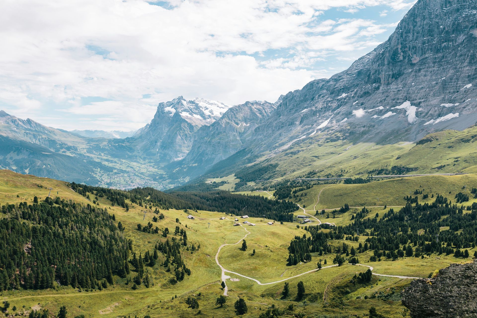 Mountain valley with a winding river, green meadows, pine trees, and rocky peaks under a cloudy sky