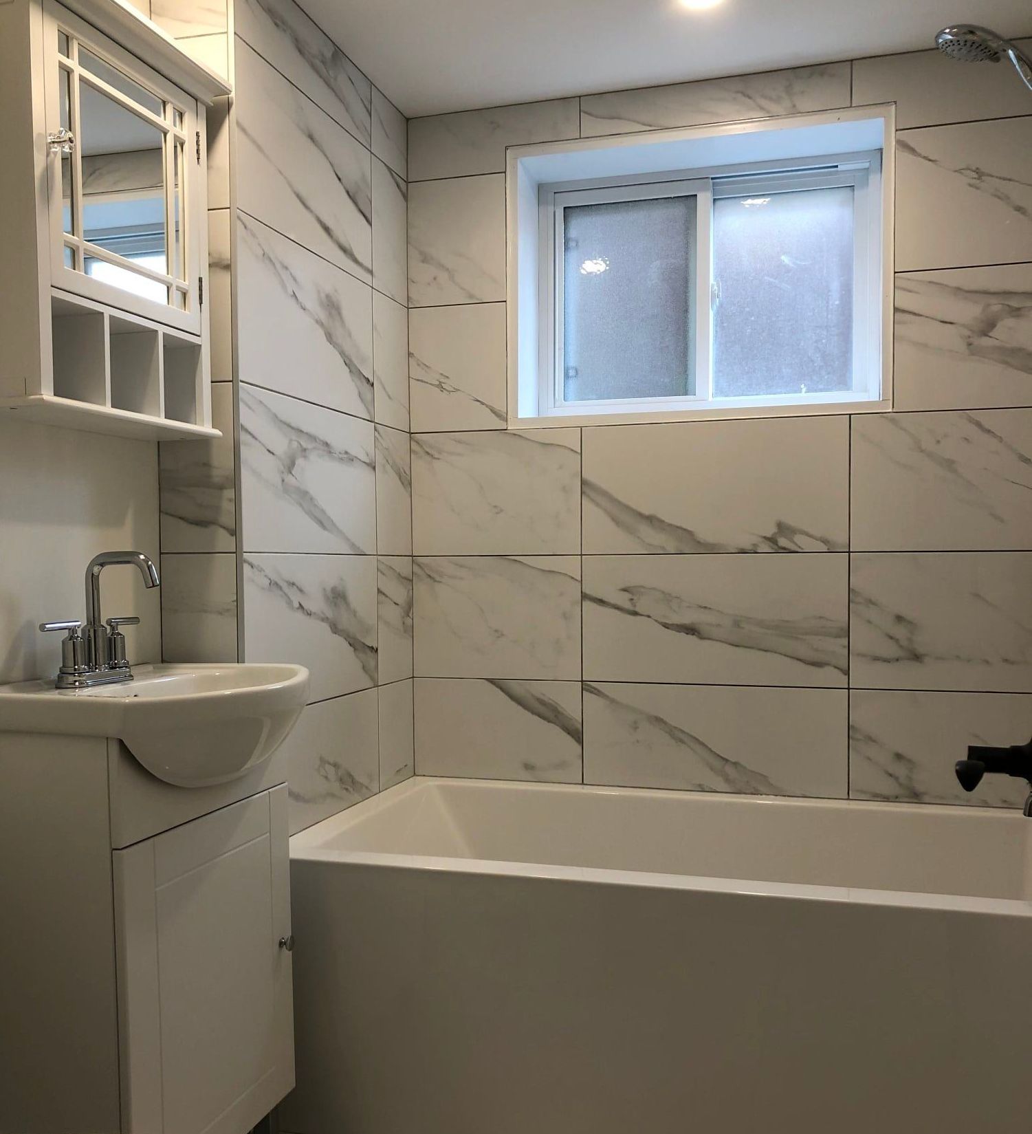 Salle de bains avec carrelage blanc imitation marbre, petite baignoire, lavabo et armoire à pharmacie avec fenêtre.