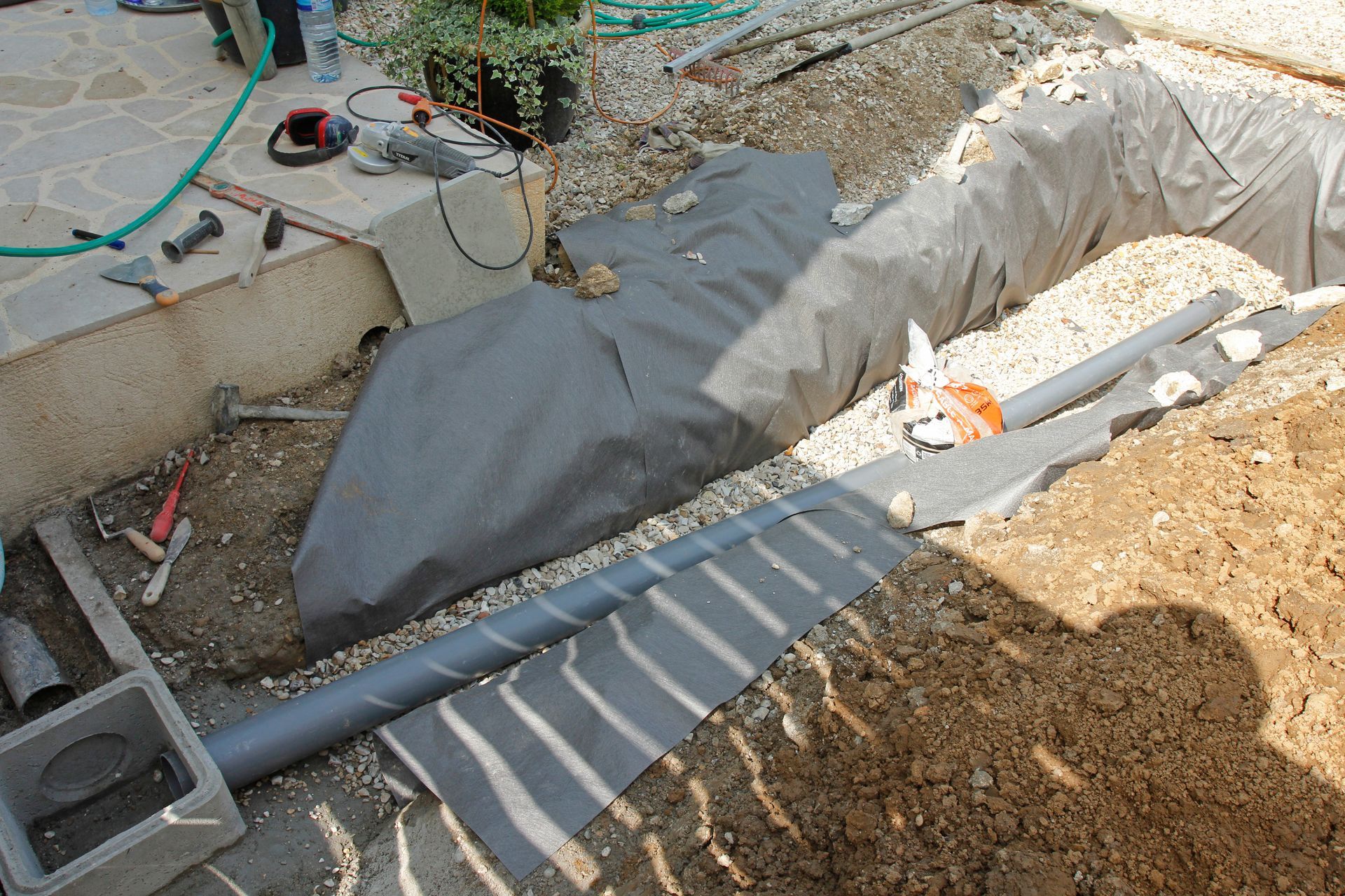 Installation d'un tuyau de drainage dans une tranchée, avec géotextile gris et gravier, près d'une terrasse en béton.
