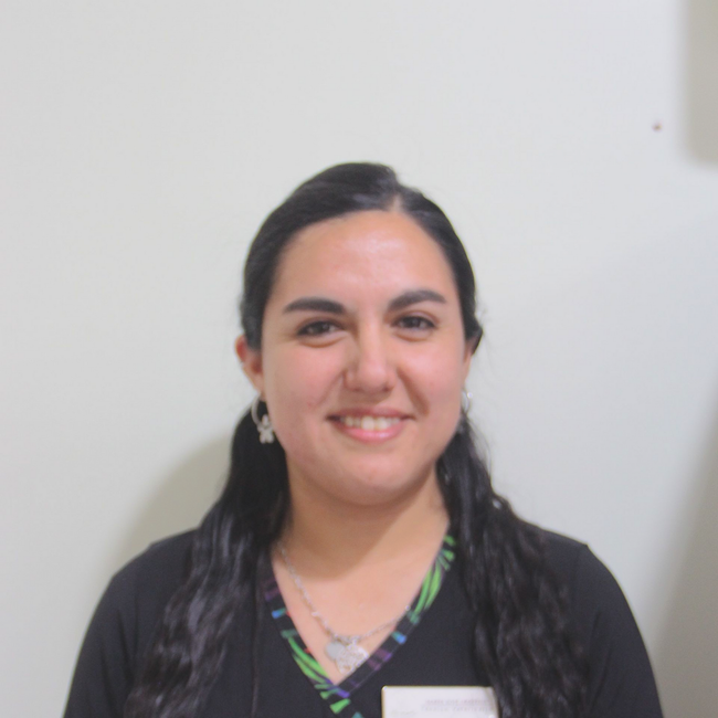 Mujer sonriendo frente a una pared blanca lisa, con una blusa oscura con cuello ribeteado en verde.