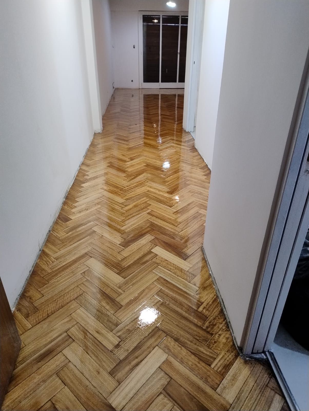 Pasillo estrecho con suelo de madera brillante en espiga y paredes blancas, que conduce a una puerta de cristal al final.