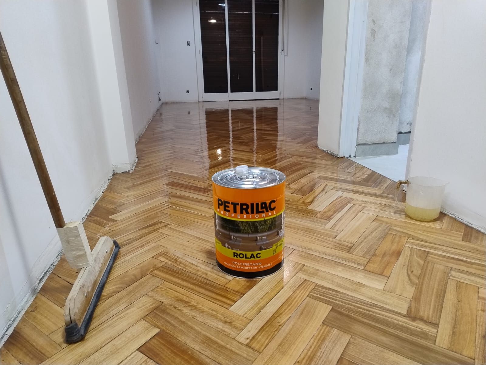 Una lata de pintura naranja sobre un suelo de madera en espiga en un pasillo, con una escoba y el acabado aún húmedo cerca.