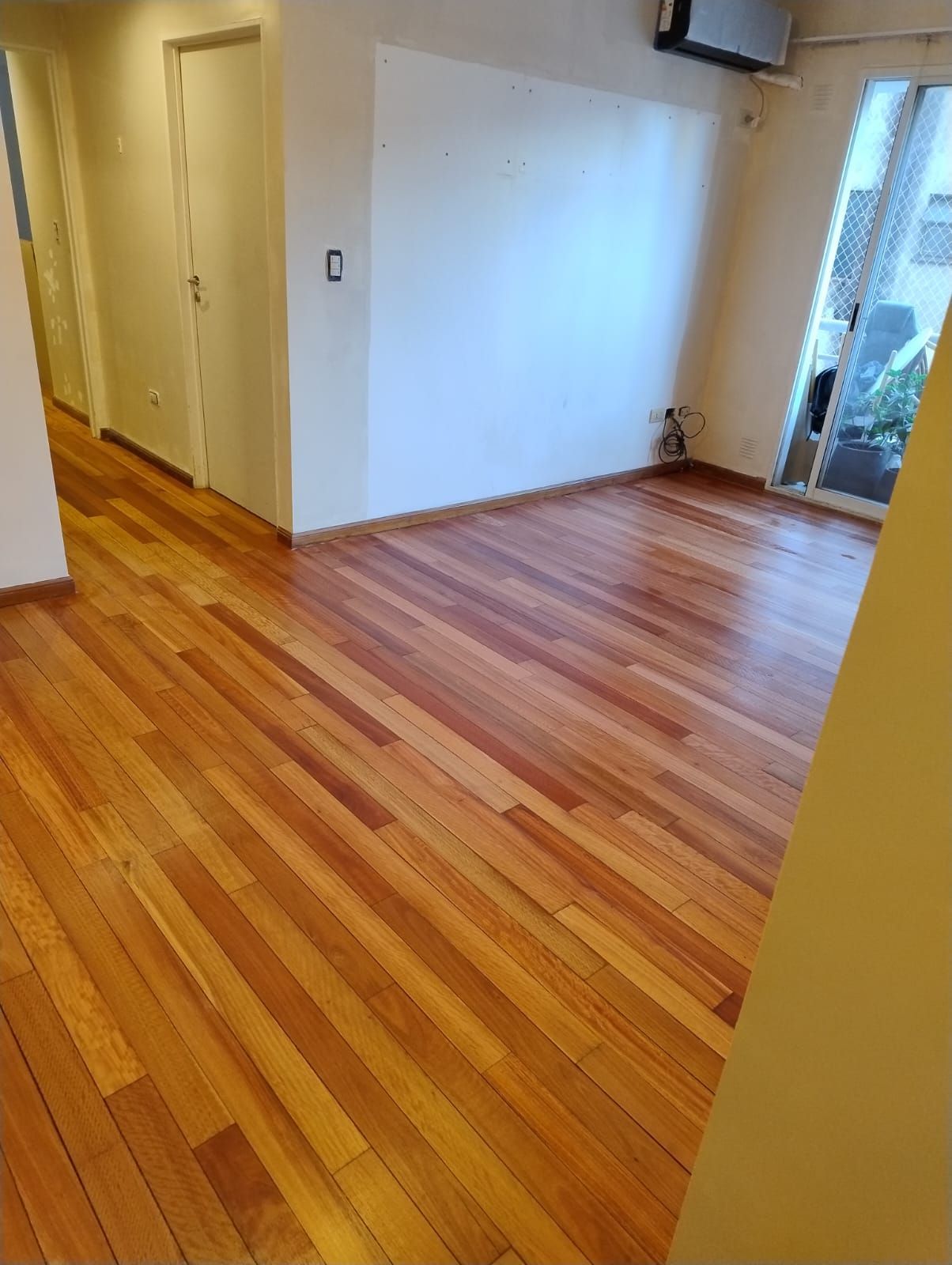 Habitación vacía con suelos de madera pulida, paredes blancas, pasillo y puerta corredera de cristal.