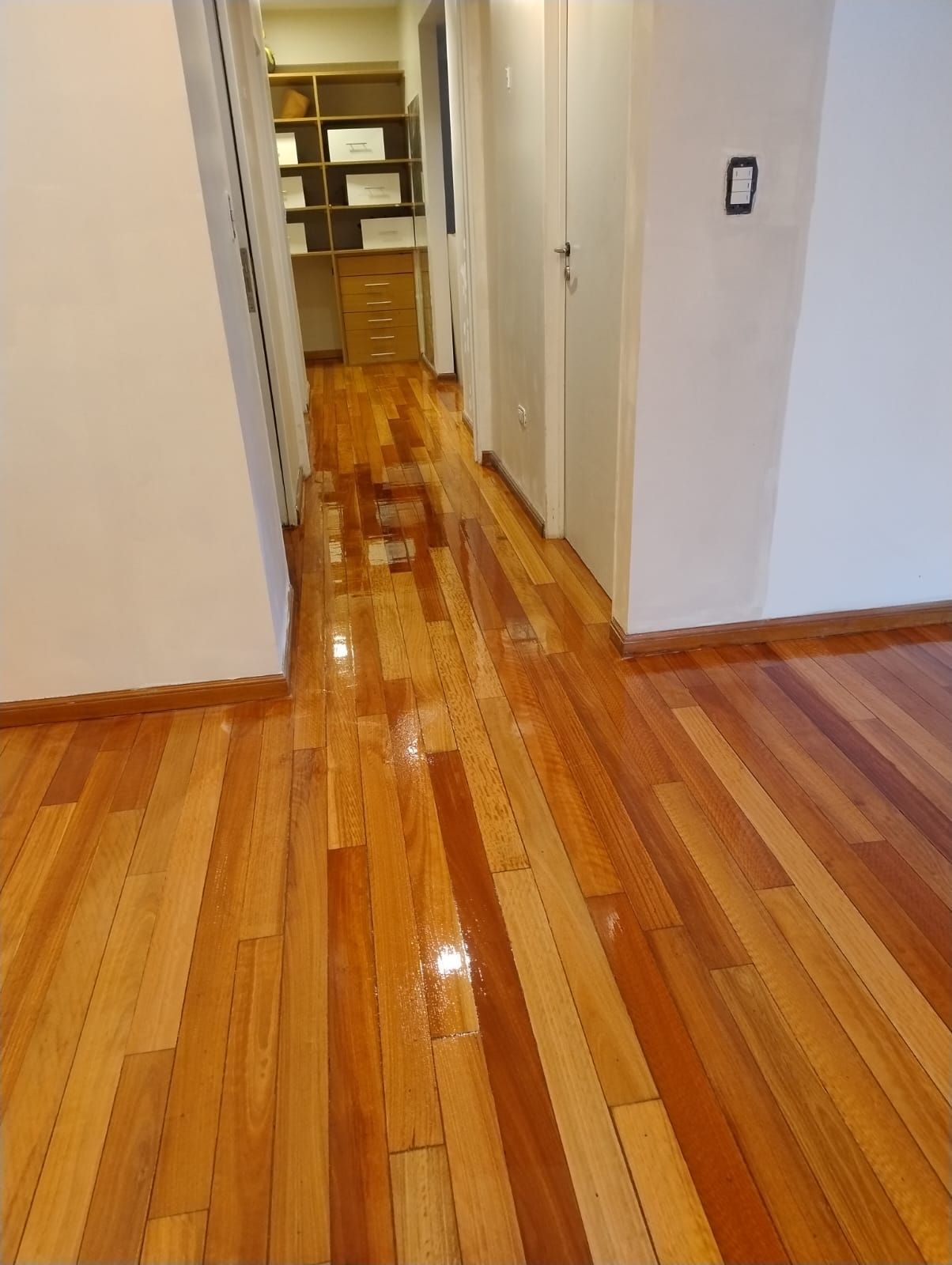Pasillo de madera pulida con paredes blancas y una puerta que conduce a una habitación con estantes.