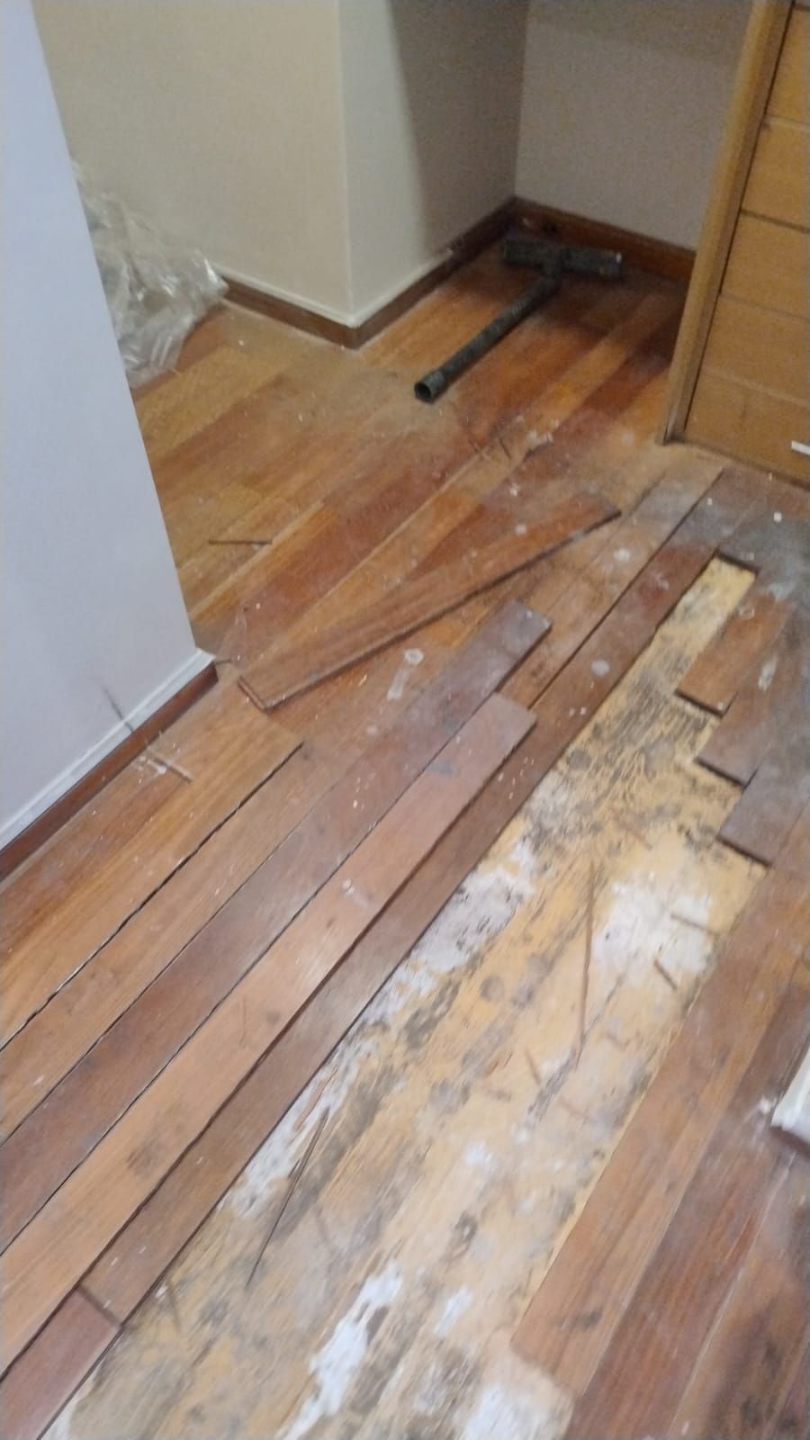 Suelo de madera noble dañado, con tablas sueltas y rotas, en la esquina de un pasillo cerca de una pared y un armario.