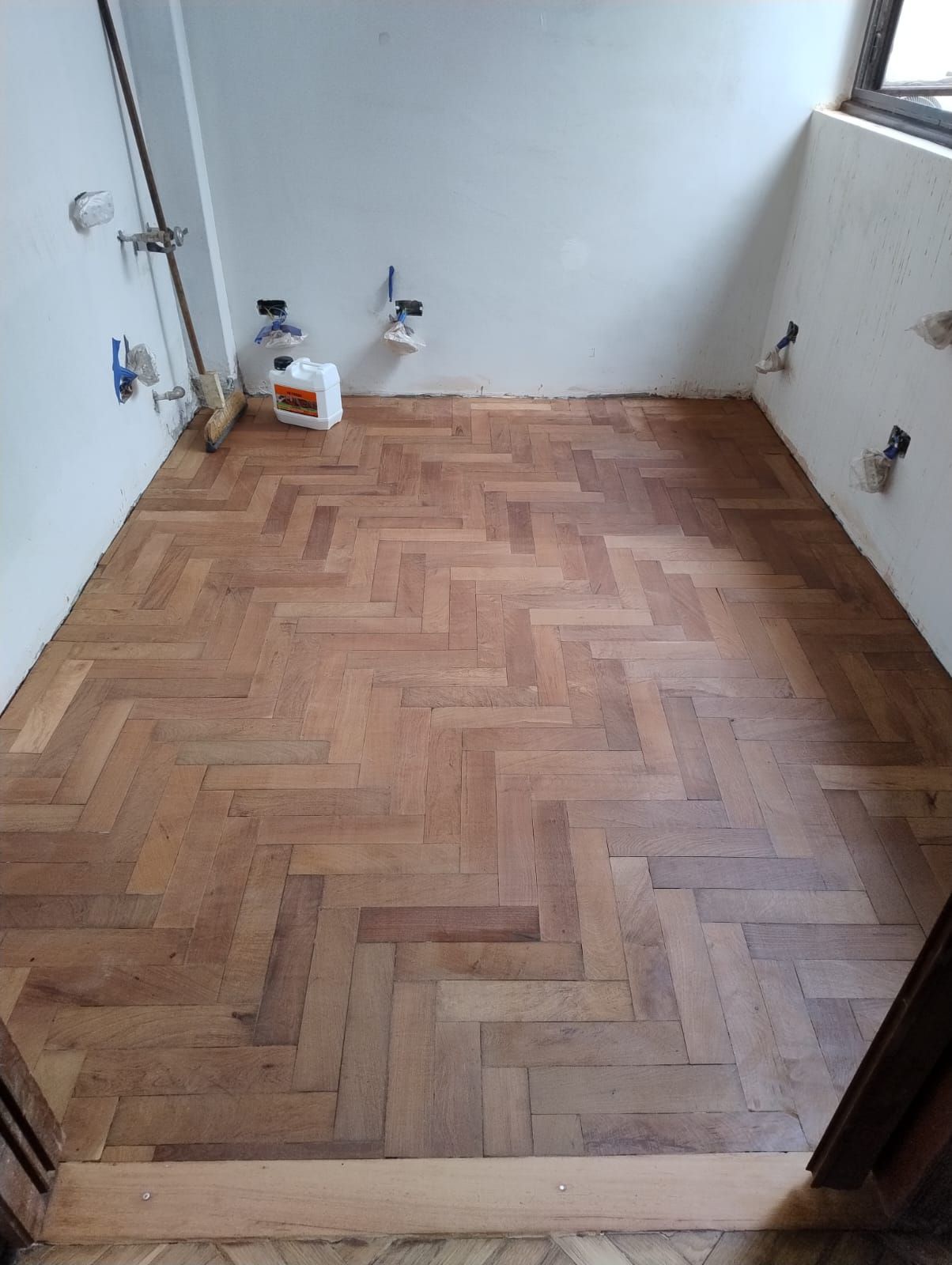 Habitación pequeña y vacía con suelo de madera en espiga, paredes blancas y accesorios de fontanería cerca de las esquinas.