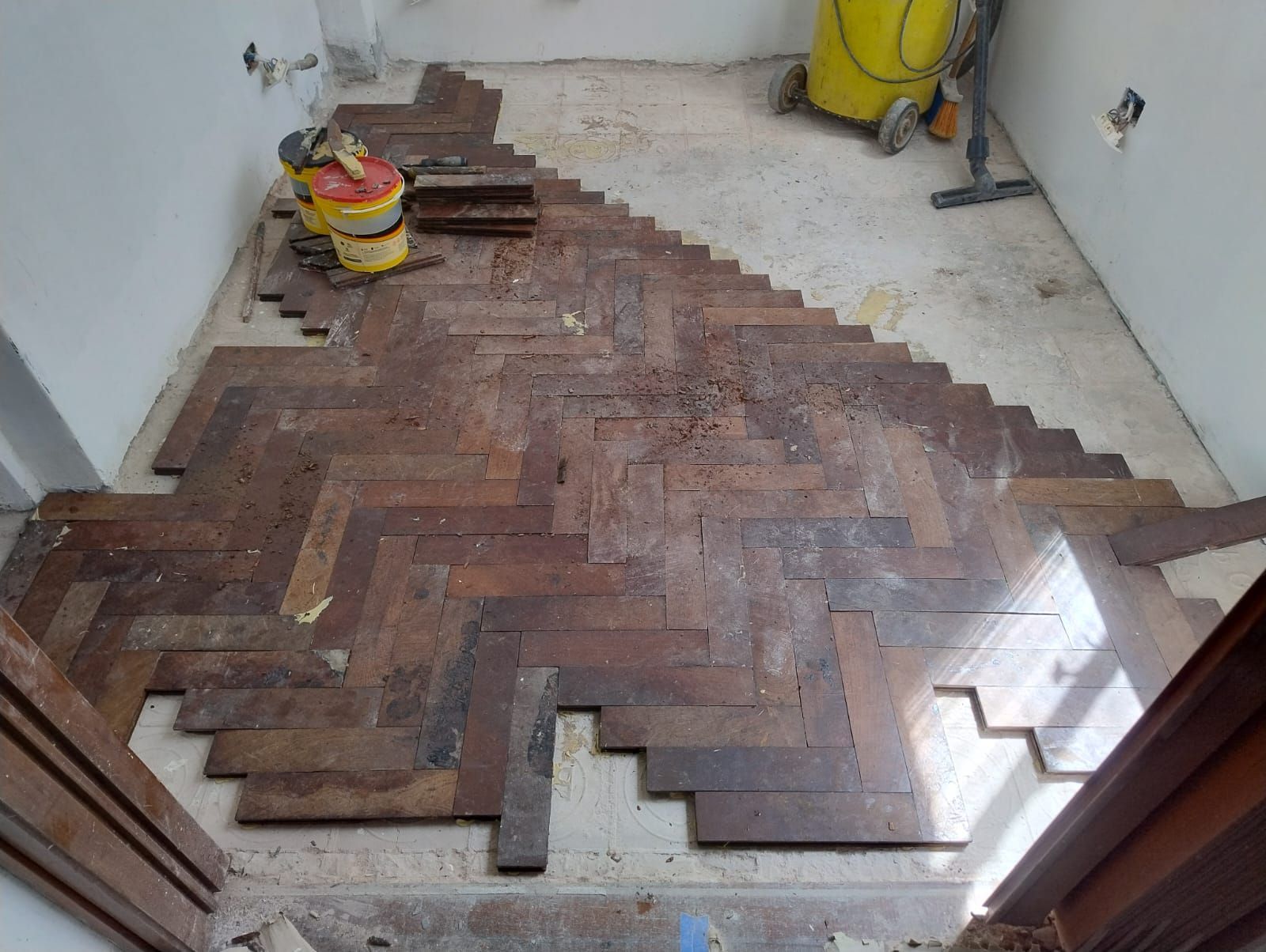En una habitación hay un suelo de madera en espiga parcialmente colocado, con herramientas y suministros cerca de la pared.