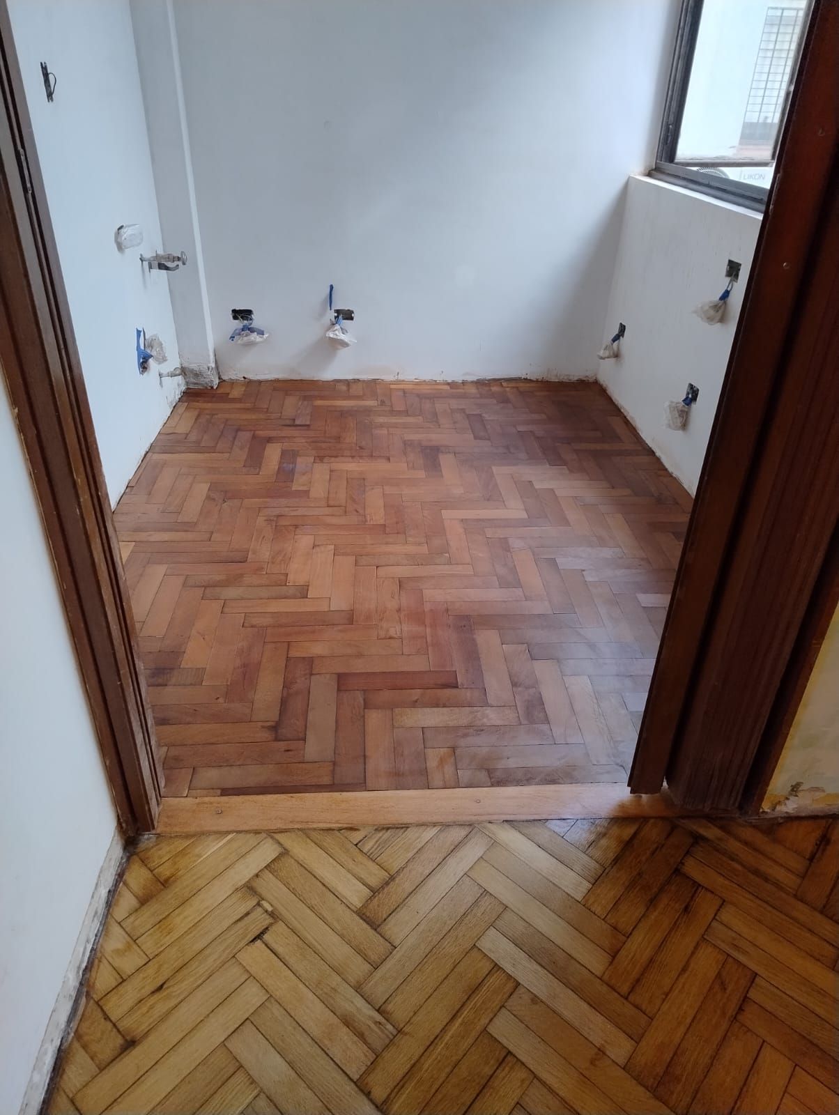 Habitación pequeña y vacía con suelo de madera en espiga y paredes blancas, vista a través de una puerta.