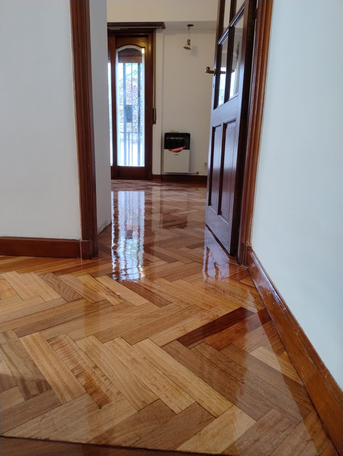 Pasillo con suelo de parqué pulido, paredes blancas y una puerta abierta que da a una habitación luminosa.