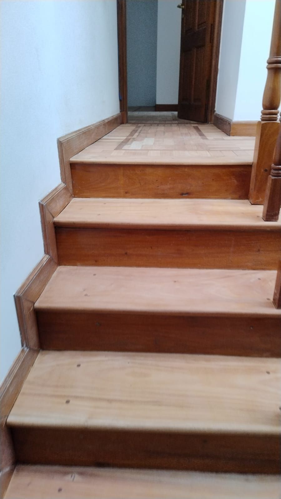Escalera de madera con varios peldaños que conduce a un pasillo, junto a una pared blanca y una barandilla.