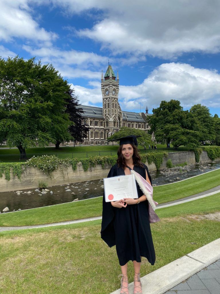 A woman in a graduation cap and gown is holding a certificate in front of a building.