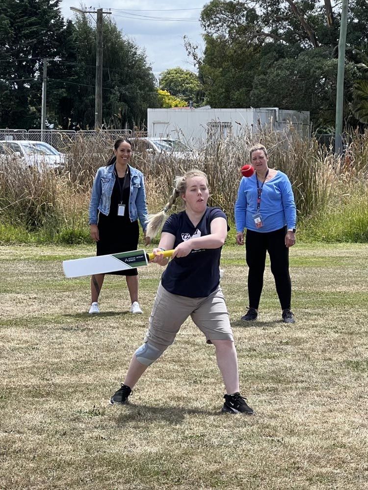 A woman is swinging a cricket bat in a field while two women watch.