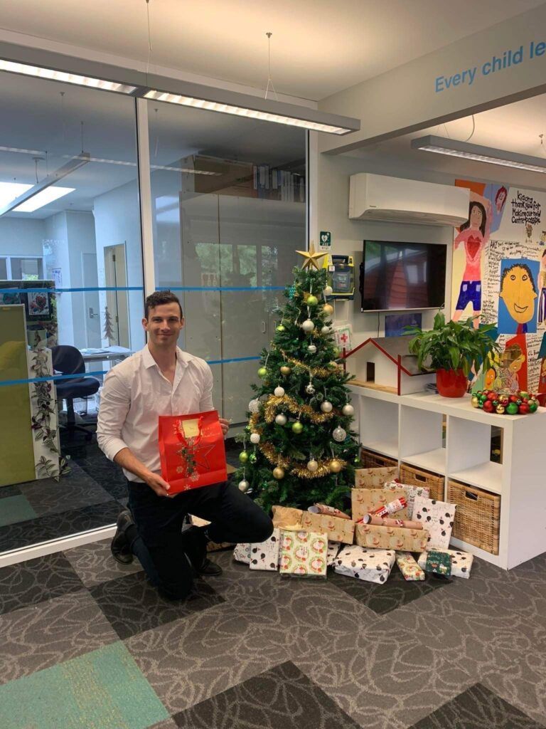 A man is kneeling down in front of a christmas tree holding a gift box.