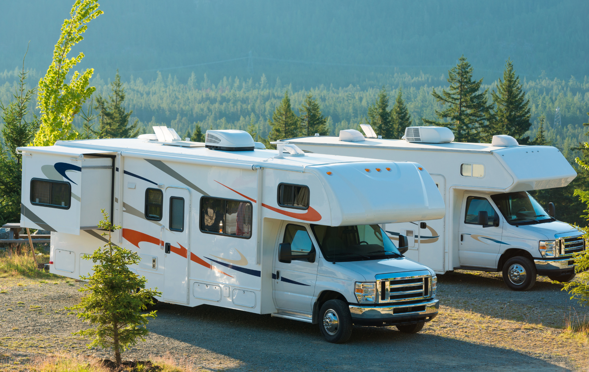 Two white RVs parked outdoors in front of a forest with a mountain backdrop.