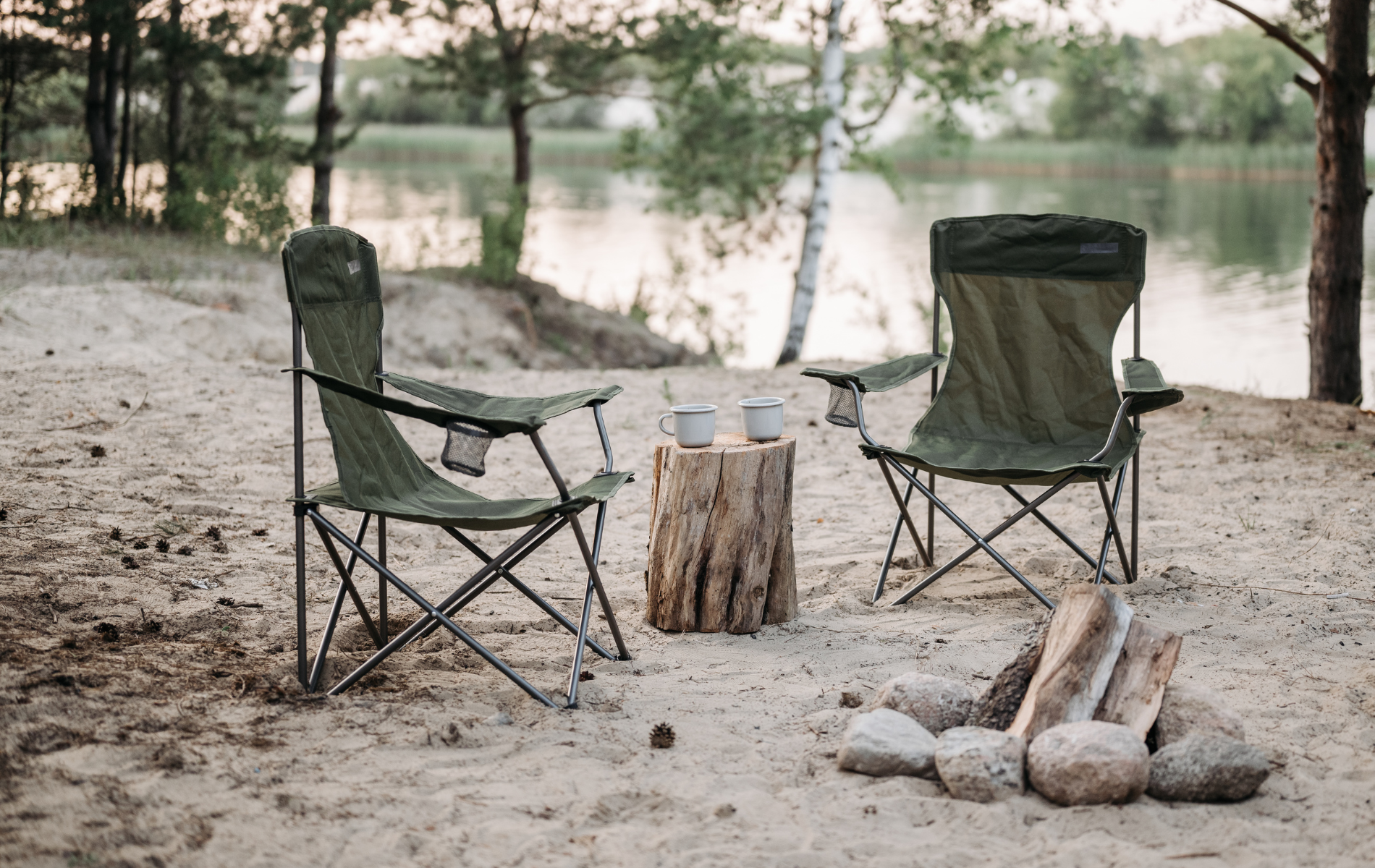 Two folding green camping chairs face a lake. A wooden stump with two cups sits between them, next to a fire pit.