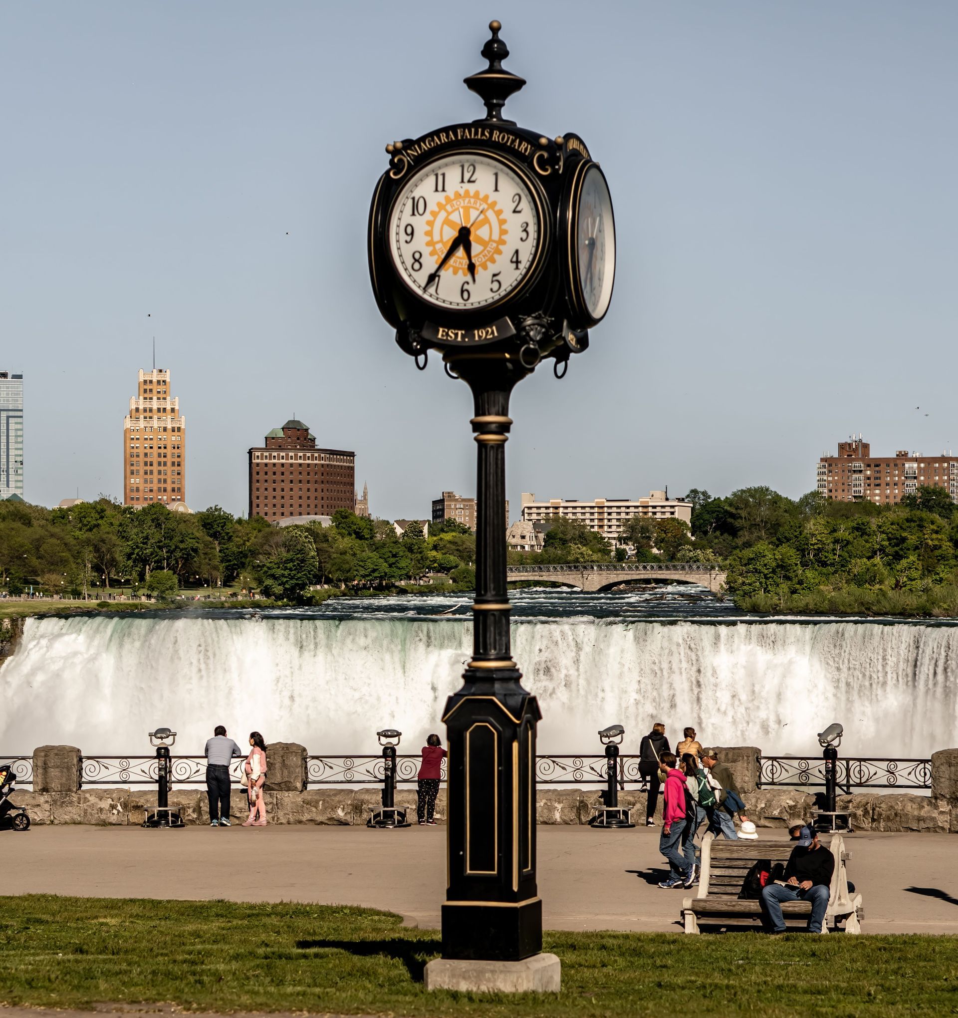 Vintage clock on a park lamppost with people, waterfall, and city skyline in the background