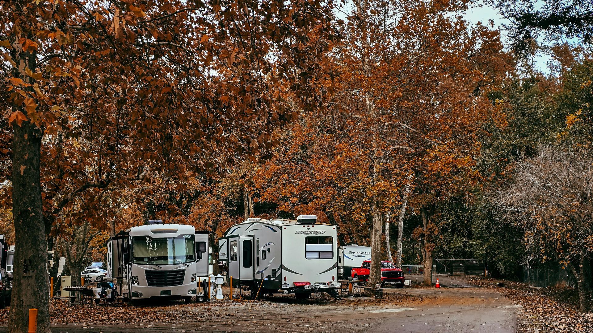Two RVs parked in a wooded campground with autumn leaves and a dirt path.