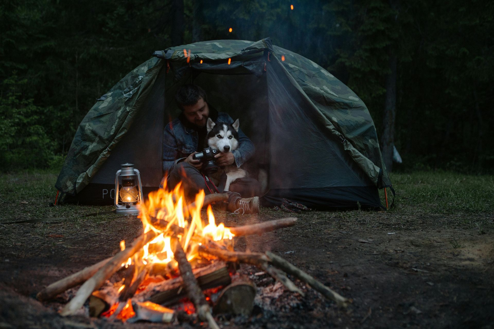 Man and husky dog camping near a campfire in a forest at dusk.