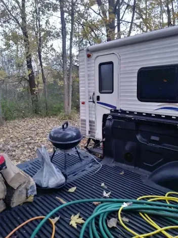 Black truck bed with a camper, grill, firewood, and hoses, parked near trees.