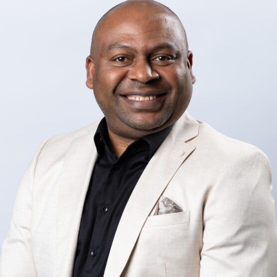 Man in cream blazer and black shirt, smiling against a gray background.