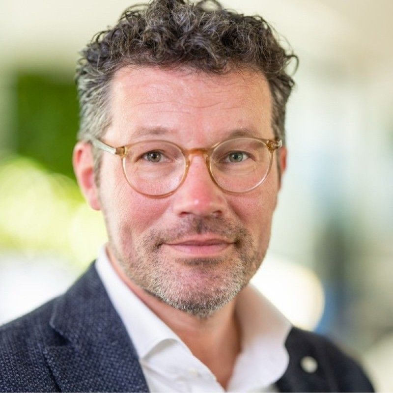 Een man met bril, blazer en wit overhemd glimlacht, binnen.