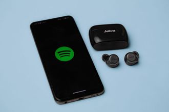 Smartphone with Spotify logo beside black wireless earbuds and charging case on a light blue background