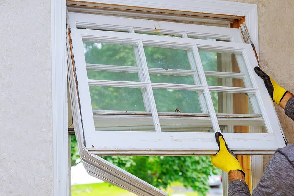 Hands installing a white window frame into a wall opening with yellow gloves