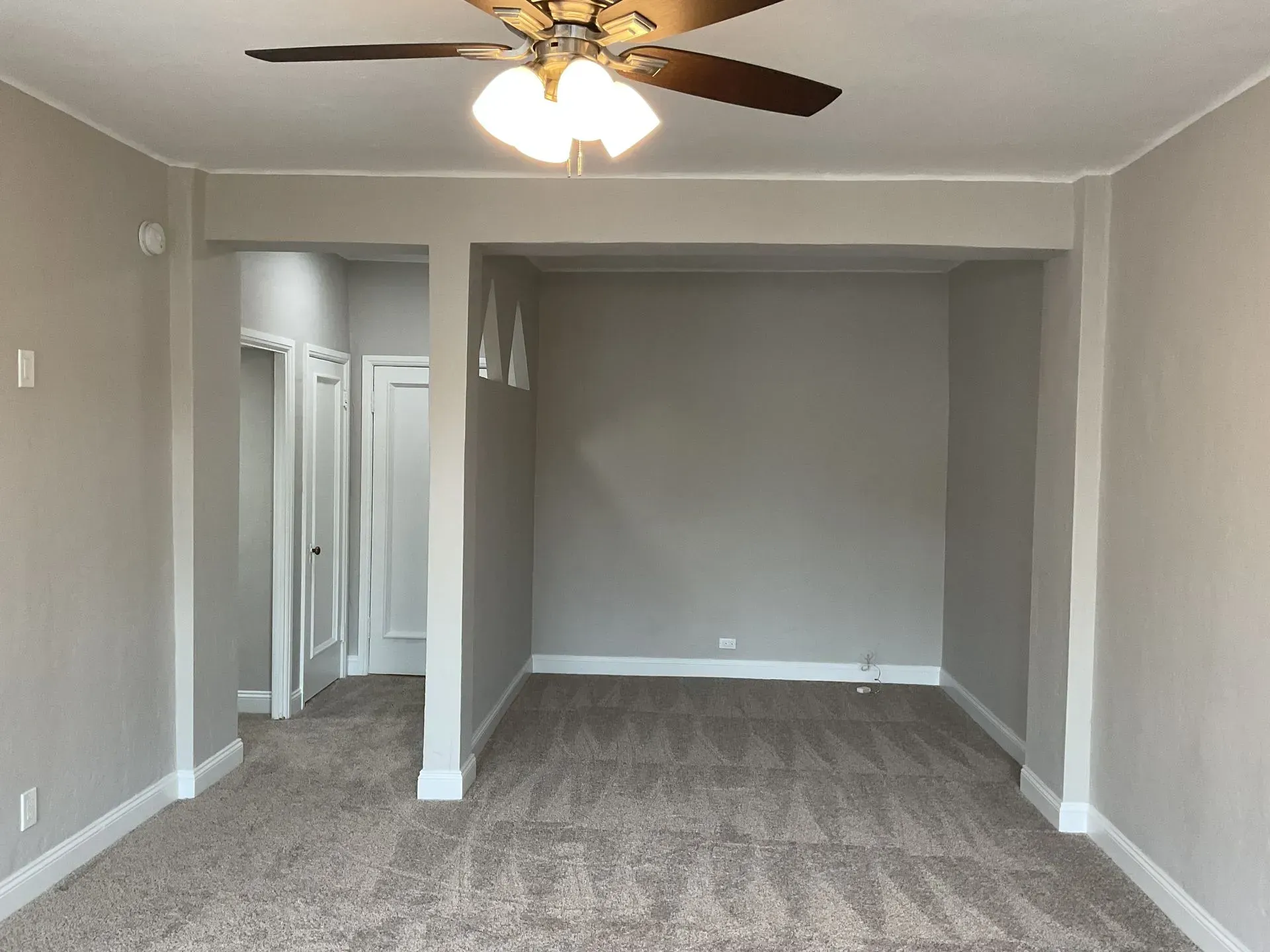Empty room with gray walls, tan carpet, and white trim. Ceiling fan. Doorways on the left and back.